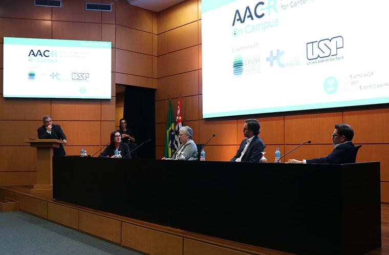 A conference hall with a stage that has a podium to left and a long black desk. There are screens both above the podium and the desk with logos for AACR on Campus, University of São Paulo, and the Institute of Cancer of São Paulo. At the podium is Roger Chammas, MD, PhD and seated at the desk is Renata Pasqualini, PhD; Patricia M. LoRusso, DO, PhD (hc), FAACR; Thomas U. Marron, MD, PhD; and Luis A. Diaz Jr., MD, FAACR.